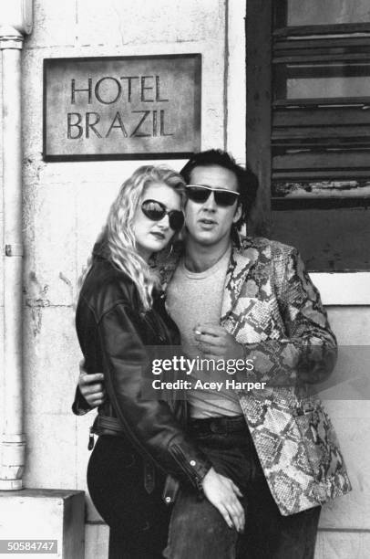 Actor Nicolas Cage wearing a snakeskin sport coat, posing w. Actress Laura Dern, both wearing sunglasses, in front of Hotel Brazil on location during...