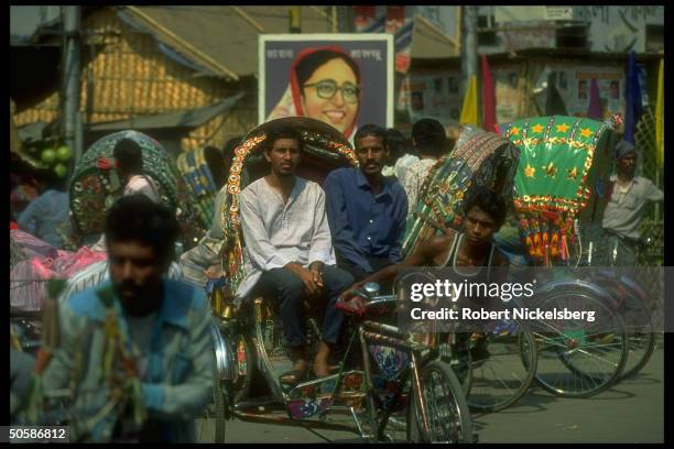 Passengers riding in bicycle- drawn taxi amid traffic passing election campaign poster bearing image of Awami League cand. Sheik Hasina Wazed.