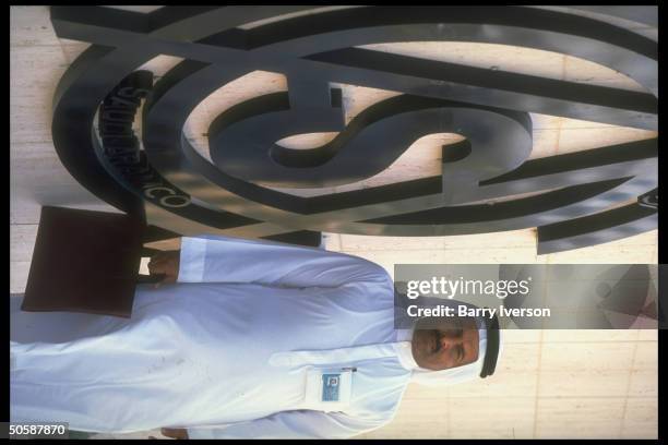 Solemn-faced, ID badge-wearing & briefcase-carrying Saudi businessman-type by Saudi Aramco logo-bearing bldg. Facade .