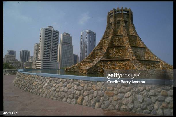 Elaborate stone fountain, w. Castle turret-like shape gracing corniche area, w. Office towers looming beyond.