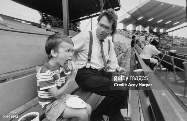 Mike Veeck, pres. Of Miracle, a minor-league baseball team, holding hot dog on a plate as he watches his 5-yr-old son William Night Train take a bite...