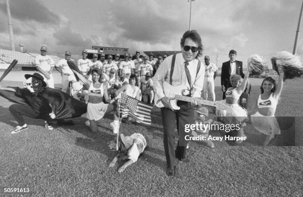 Mike Veeck, pres. Of Miracle, a minor-league baseball team, playing electric guitar as he poses in front of 4 cheerleaders & two dozen Miracle...
