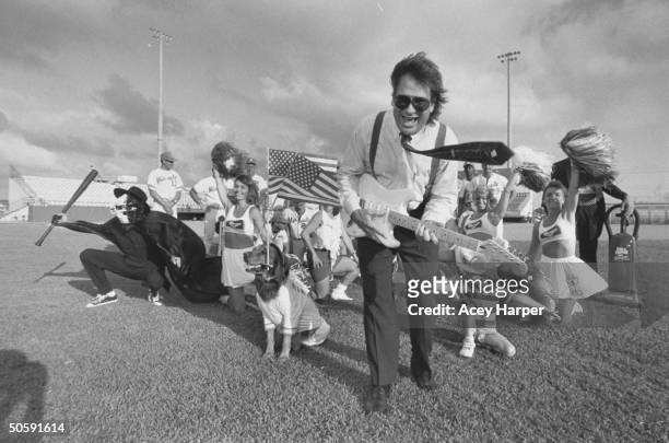 Mike Veeck, pres. Of Miracle, a minor-league baseball team, playing electric guitar as he poses in front of 4 cheerleaders & two dozen Miracle...