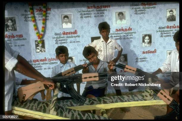 Boys on mil-theme seesaws framed by pics & birth/death dates of prob. Young victims of LTTE Tamil Tiger secessionist war, at playground in...