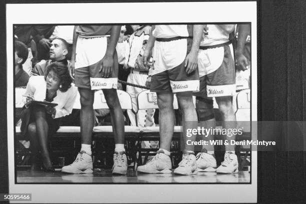 Bernadette Locke-Mattox, asst. Coach at Univ. Of KY tensely holding clipboard while sitting on bench straining to look out from nearby legs of 3...