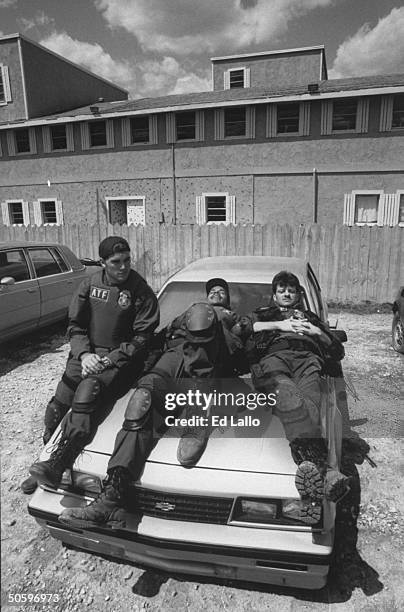 Movie extras Kit Clark, Patrick Allmano & Bobby Young, all clad in ATF agent's combat garb as they stretch out on the hood of a car in front of set...