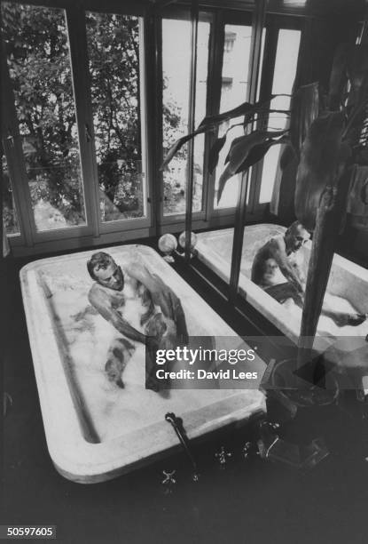 Fashion designer Giorgio Armani taking a bubble bath in his large marble bathtub next to mirrored wall in bathroom at home.