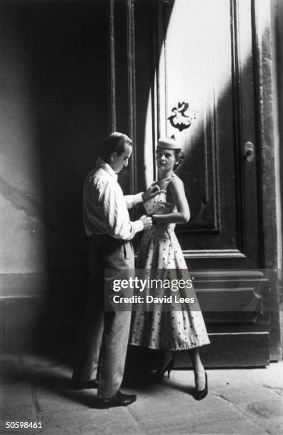 Italian fashion and fabric designer Emilio Pucci standing in open doorway helping model fit into dress he designed.