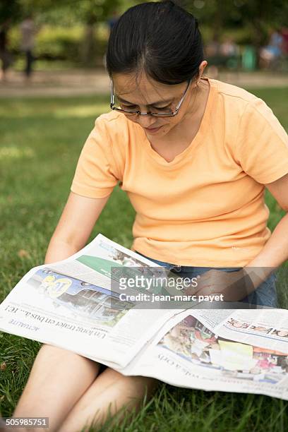 French woman of asian origin sat on the grass in a park reading the newspaper " The Parisian " in the afternoon. Model release available