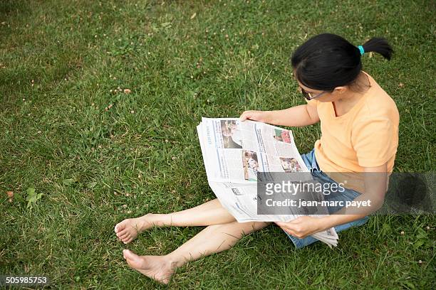 French woman of asian origin sat on the grass in a park reading the newspaper " The Parisian " in the afternoon near Paris in France. Model release...