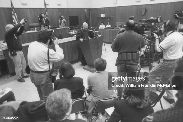 Judge Larry Gram standing at podium in front of the bench in his courtroom, speaking to the reporters & cameramen surrounding him at press conference...