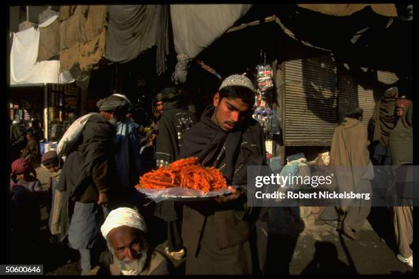 Main street market stalls framing youthful vendor carrying confection-heaped tray in Kandahar, Afghanistan.