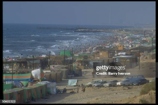 Beach tents & plastic-covered cabanas framed by Gazans cooling off in water, enjoying day at beach in Palestinian autonomous Gaza.