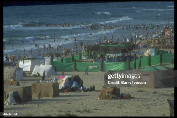 Beach tents & plastic-covered cabanas framed by Gazans cooling off in water, enjoying day at beach in Palestinian autonomous Gaza.