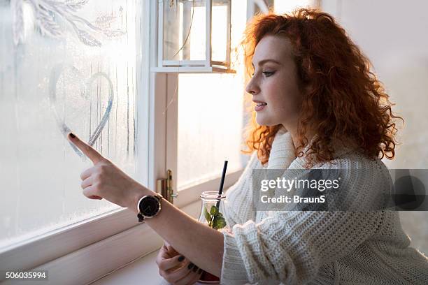 young smiling woman drawing a heart on a window. - condensation stock pictures, royalty-free photos & images