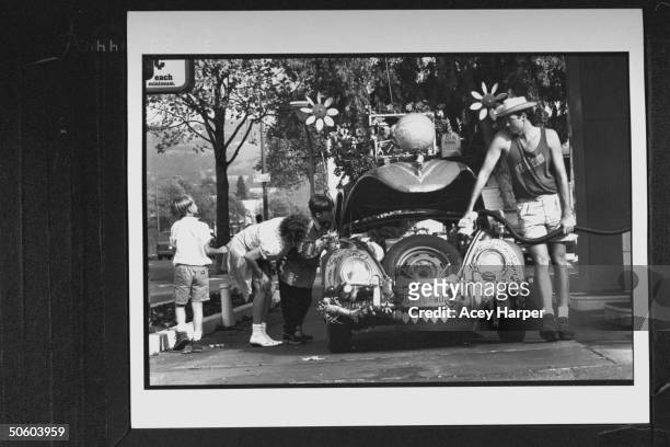 Auto artist/filmmaker Harrod Blank pumping gas into his VW Bug as Terri Sly & her 2 sons Jared & Josh Conley inspect the car adorned w. A globe hood...