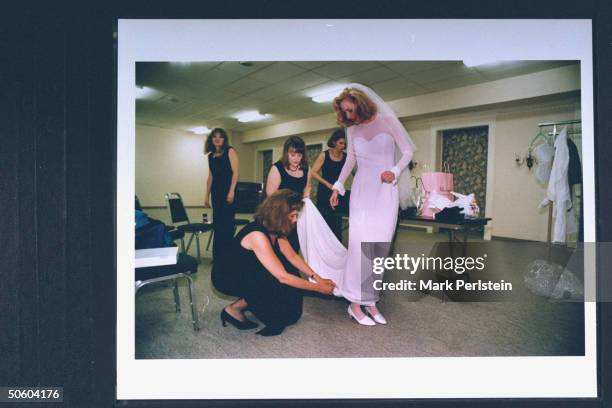 Law student/bride Cindy Dyer , wearing a simple white wedding dress, as 1 of 4 bridesmaids in full-length black sheath gowns fixes Dyer's hem on the...