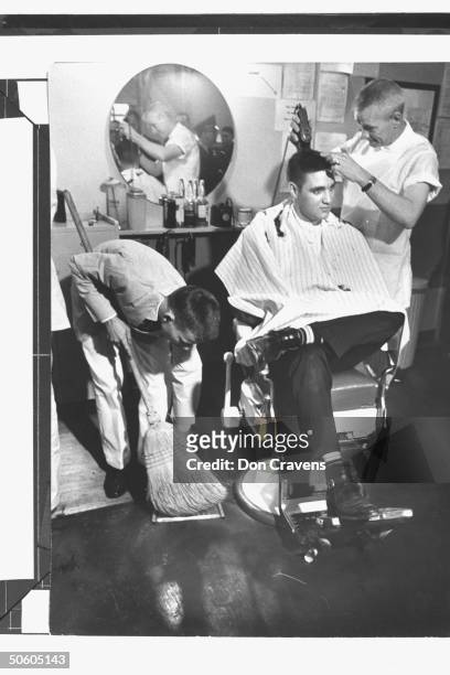 Singer/Army Pvt. Elvis Presley sitting in chair while Army barber James Peterson brush-cuts his famous mane as man sweeps up hair off the shop's...