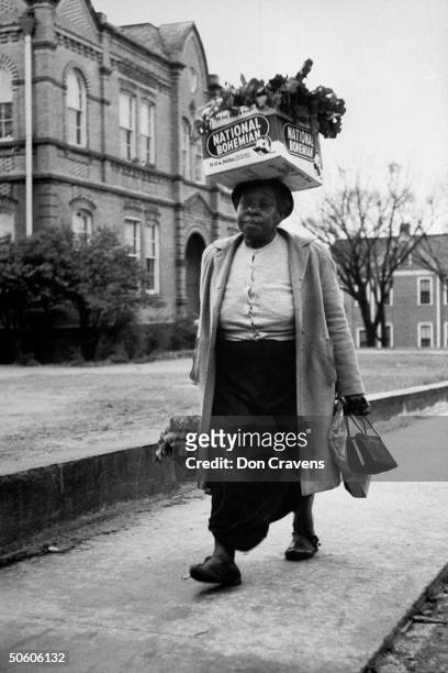 Black woman w. National Bohemian beer box loaded w. Turnip greens balanced on her head, walking on sidewalk during bus boycott protesting policy of...