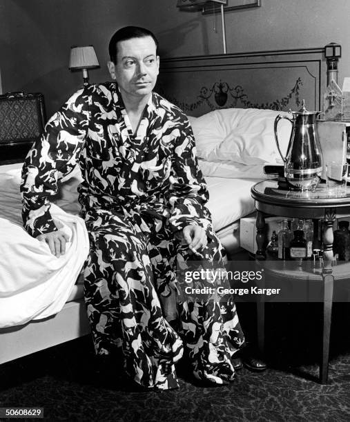 American composer Cole Porter sits on edge of bed in his room at the Ritz Hotel, a cigarette in his hand and dressed in a robe with a wild horse...