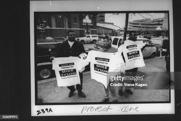 Three black Tyson fans holding up T-shirts emblazoned w. The slogan JUSTICE AND FAIRNESS FOR MIKE TYSON as they go about trying to sell the fact that...