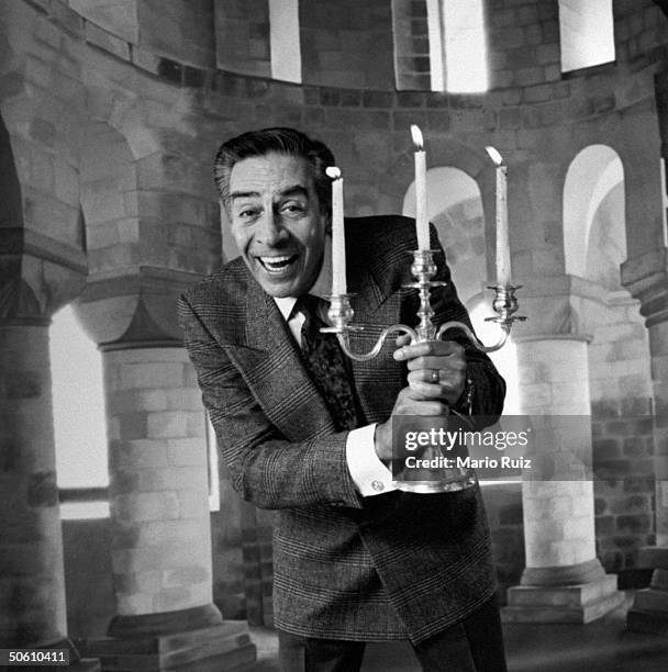 Actor Jerry Orbach holding a silver candelabra w. 3 lit candles in front of backdrop at studio, acting like the character Lumiere, the candlestick,...