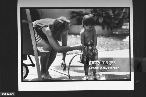 Newswoman/unwed mom Jane Wallace tickling her adopted 30-mo-old son Zach w. A paint brush as he stands shirtless holding a garden hose.