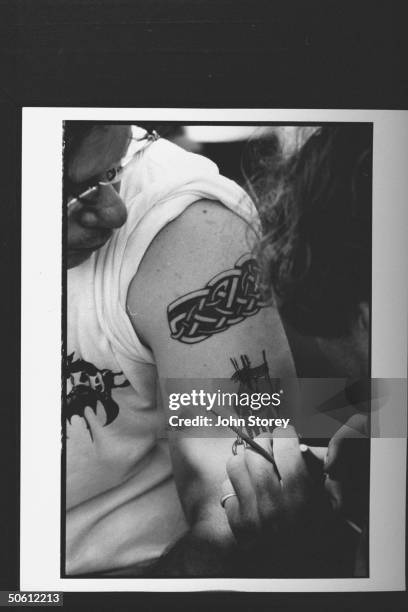 Closeup of a faux tattoo artist painting his rendition of the rope-bound ankles of a high-heeled female on the left bicep of a male concertgoer who...