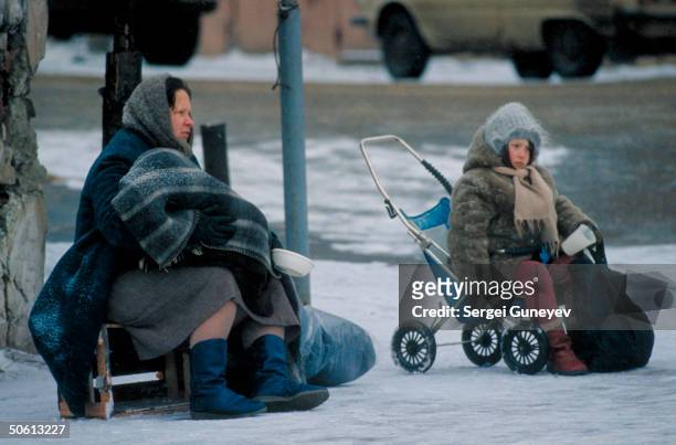 Pair of mother & daughter type beggars out in cold of winter, sitting, holding bowl & cup for solicitations, woman on crate, girl in too small...