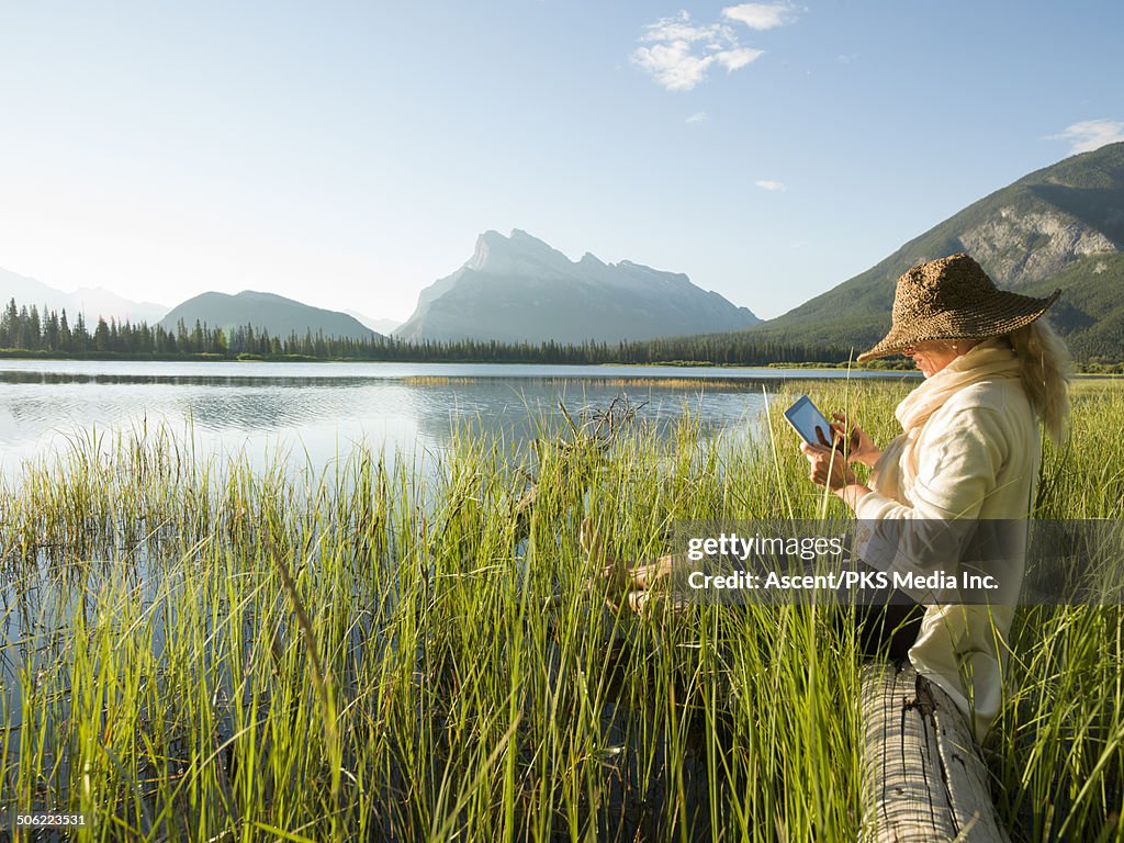 Woman relaxes on log, uses digital tablet, lake