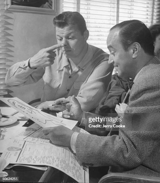 Movie dir./artist Jean Negulesco working on a caricature drawing of comedian Jack Carson who is pointing out that Jean made the nose too long, during...