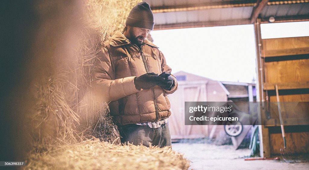 Man leaning against hay in barn looks down at phone