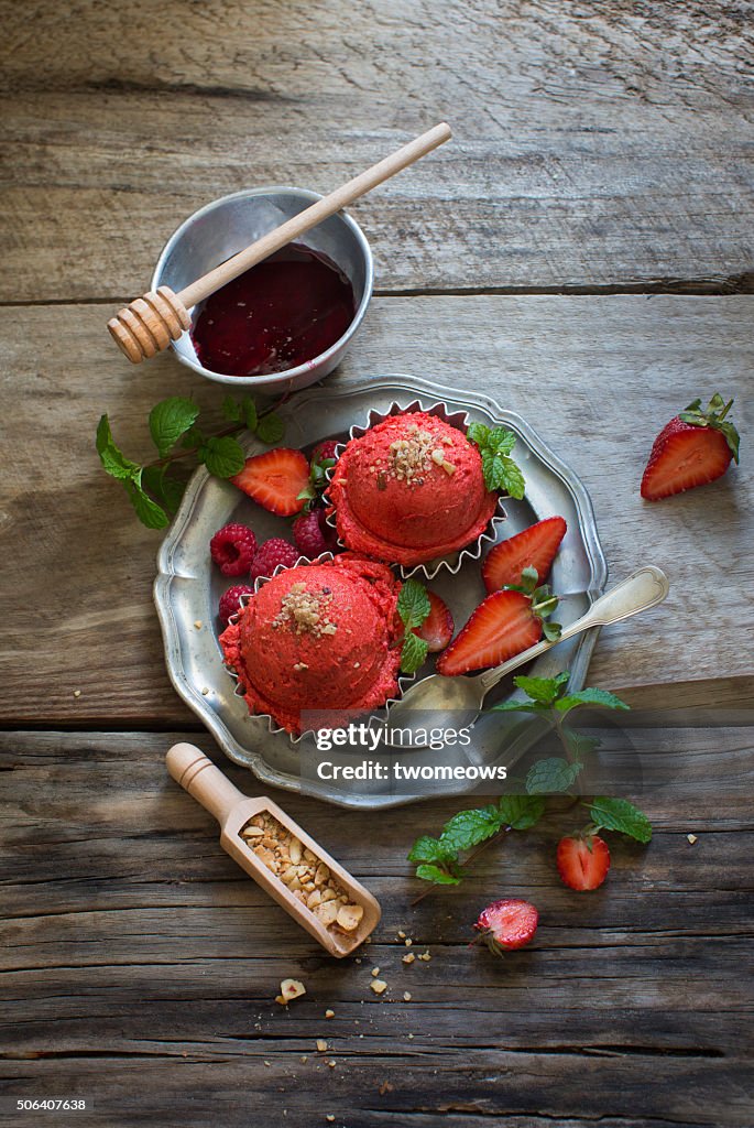 Strawberry ice cream balls with fresh berries and mint leaf served with strawberry syrup on moody rustic wooden table top.