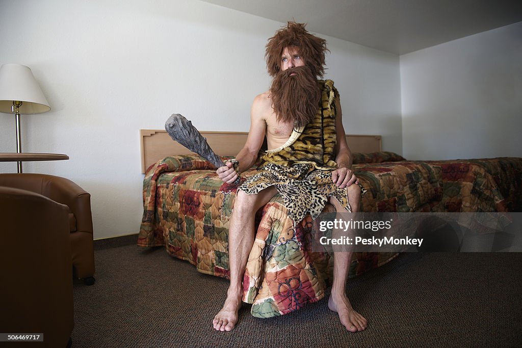 Traveling Caveman Sitting on Old Fashioned Motel Room Bed