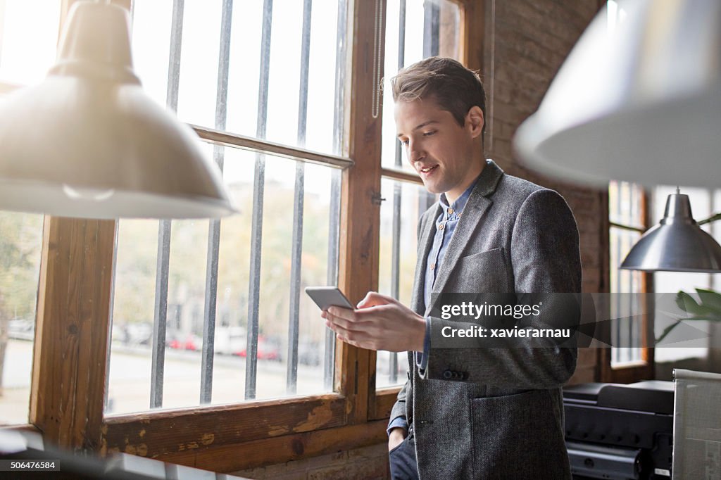 Young creative businessman at the window looking on mobile phone