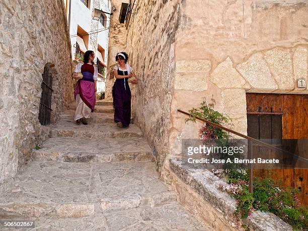 Two young women with clay pots, walking down a narrow street in old medieval neighborhood with old clothes; in a staging as lived in the past