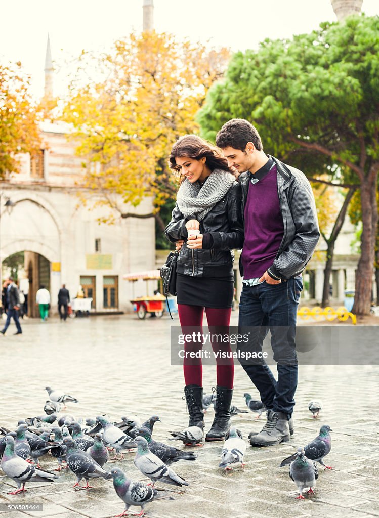 Lovely Young Couple Feeding The Birds In Town Square