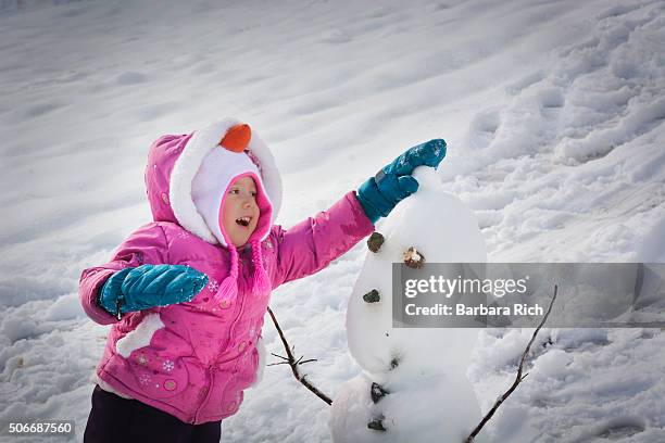 three year old blue eyed girl playing in the snow placing snowball on top snowman's head - barbara young stock pictures, royalty-free photos & images