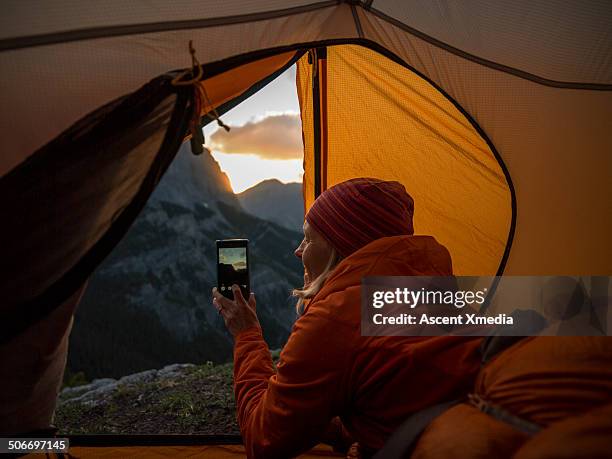 woman takes smart phone pic of sunrise, in tent - zelt stock-fotos und bilder