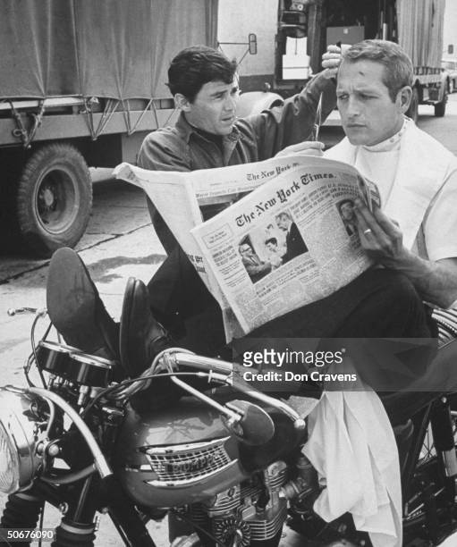 Chic barber Jay Sebring, working on actor Paul Newman's hair while he is reading the paper, sitting on motorcycle on the set of the film Moving...