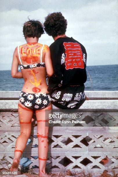 Young hippie couple, she bikini clad and painted w. Psychedelic designs, looking out at the sea during a love-in fest.