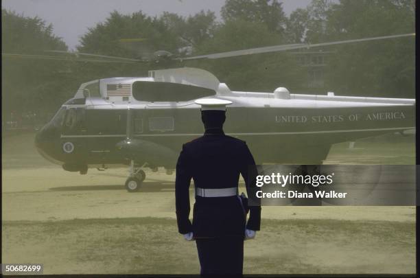 Side view of Marine One helicopter as it lands on sandy, beach-like ground, a single marine in dress uniform stands guard in foreground.