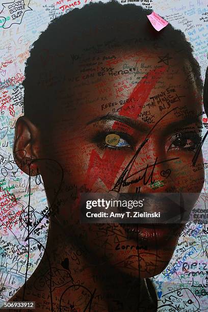 Graffiti and other writings cover a cosmetics advertisement on a wall to the left side of the flowers and other items left on a mound that continues...