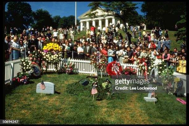 Visitors at grave site paying respects to slain President John F. Kennedy on first birthday after assassination as military guard stands by.
