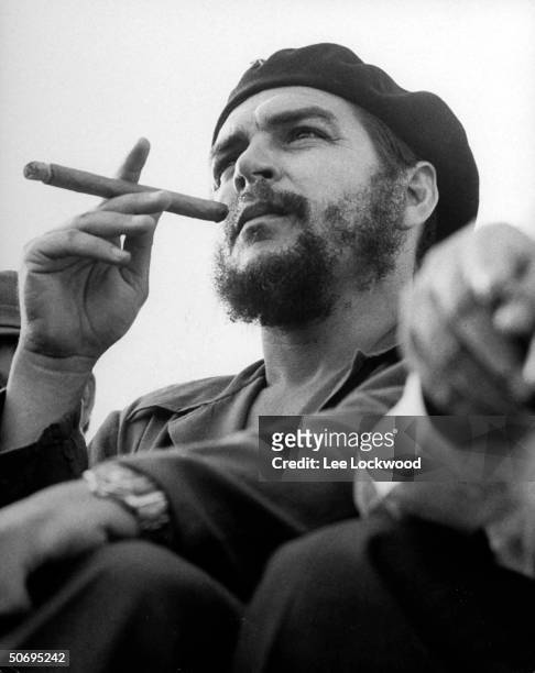 Revolutionary Che Guevara sitting on viewing stand smoking big cigar at an anniversary celebration of the Cuban revolution.