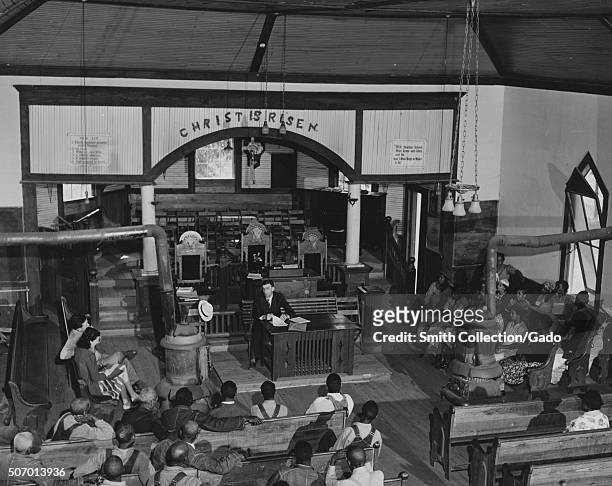 African-American Farm Security Administration borrowers sitting in a church and listening to an official, with a message above the altar reading...