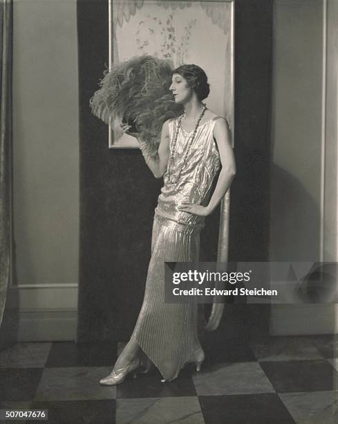 Model, Desiree Lubovska, standing with left hand on hip, holding an ostrich fan, and wearing a shimmering two-piece dress with sleeveless top and...