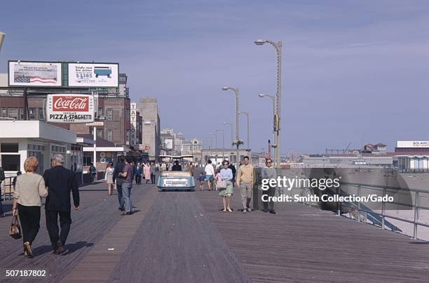 Tourists strolling on the boardwalk in Atlantic City, New Jersey, with signs advertising Coca Cola, a computer printer, and a pizza restaurant, beach...