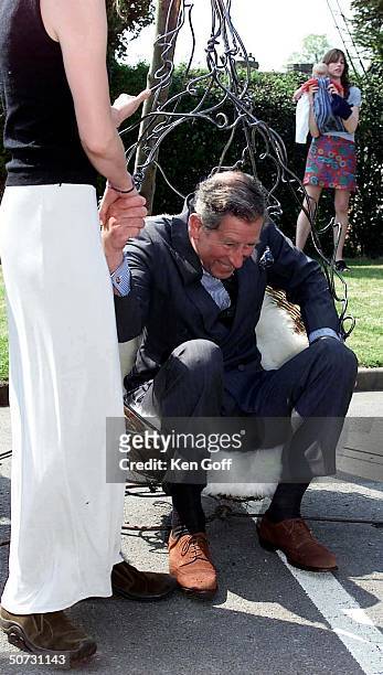 England's Prince Charles being helped out of wrought iron hanging chair suspended from 3 tree trunks made by female blacksmith during visit to meet...