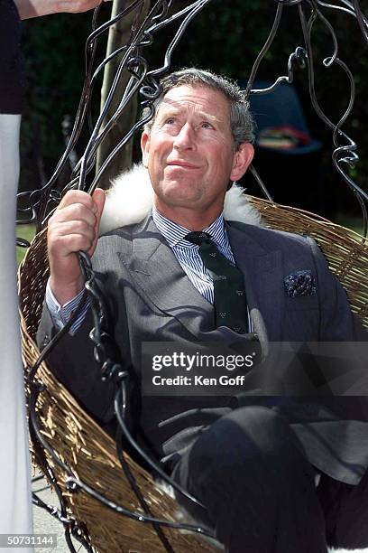 England's Prince Charles enjoying wrought iron hanging chair suspended from 3 tree trunks made by female blacksmith during visit to meet young men...